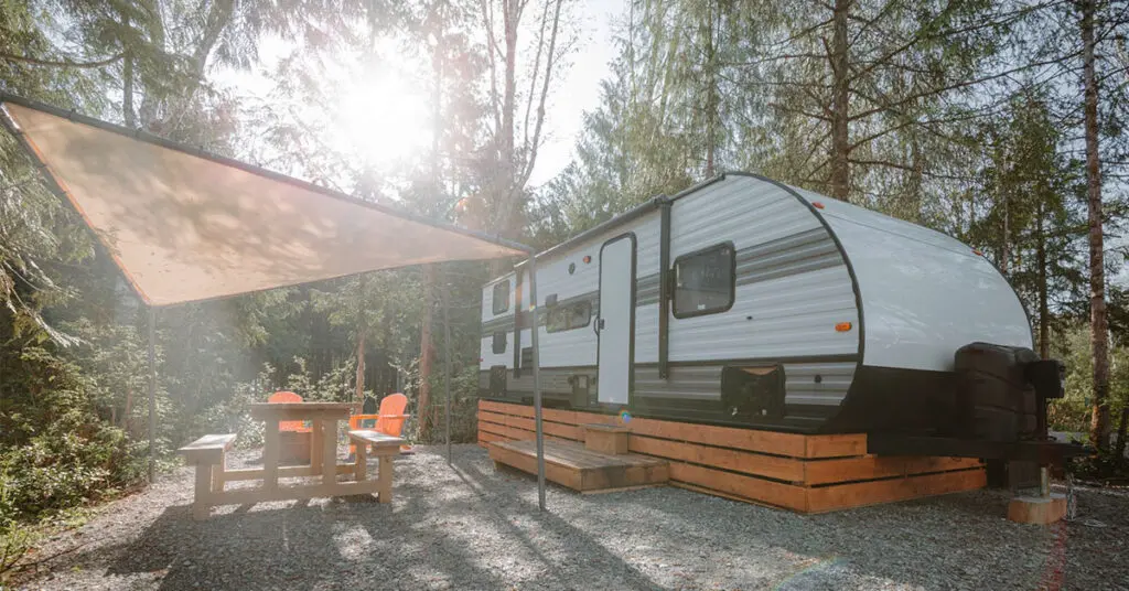 Modern travel trailer at a wooded campsite with a picnic table and sunshade.